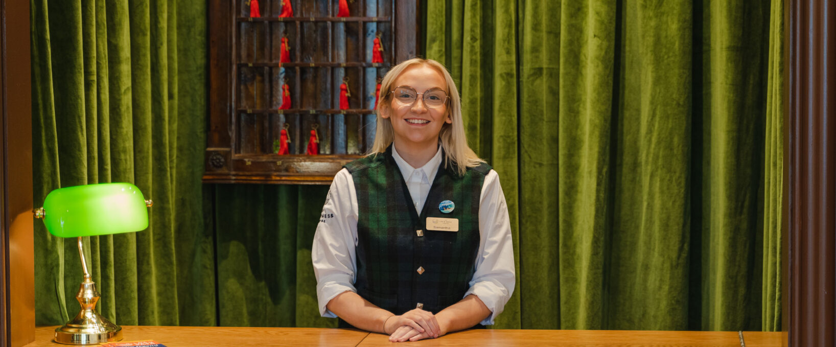 A smiling women standing at the admissions desk in Loch Ness tartan ready to welcome guests to The Loch Ness Centre